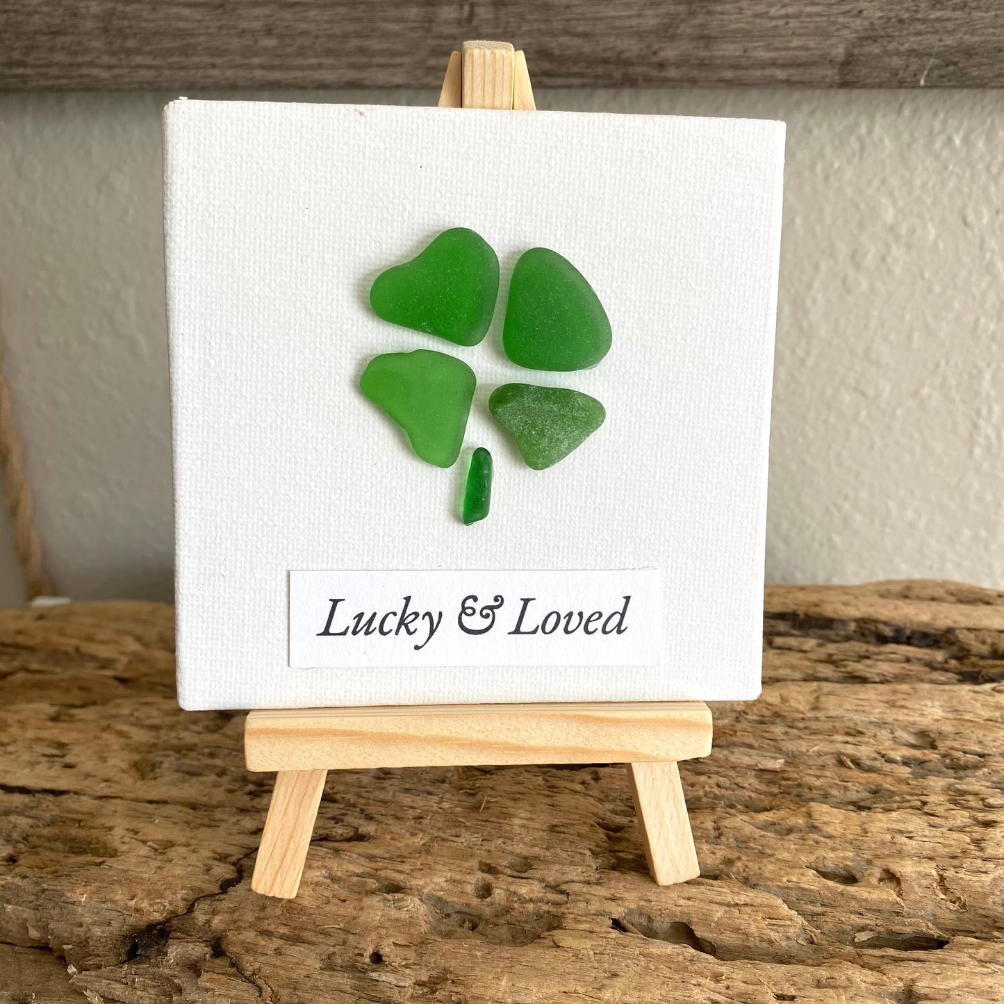 Four green sea glass pieces on a white canvas with 'Lucky & Loved' text, displayed on a wooden easel.