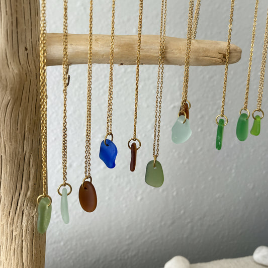 Gold sea glass necklaces with colorful pendant drops hanging on a wooden branch against a light gray background